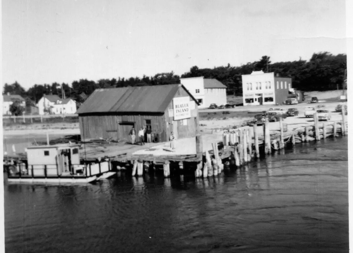 A wooden building stands on a pier beside a small boat, with parked cars and buildings visible beyond the water.