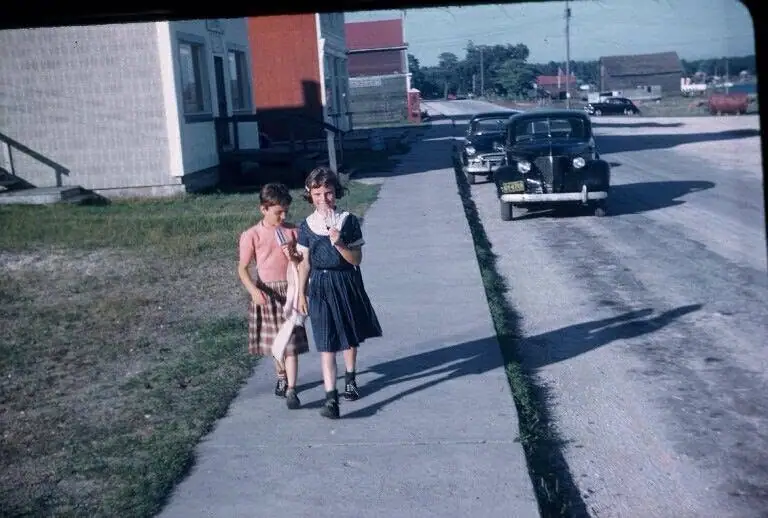 Two girls walk along a sidewalk beside parked vintage cars and low-rise buildings.