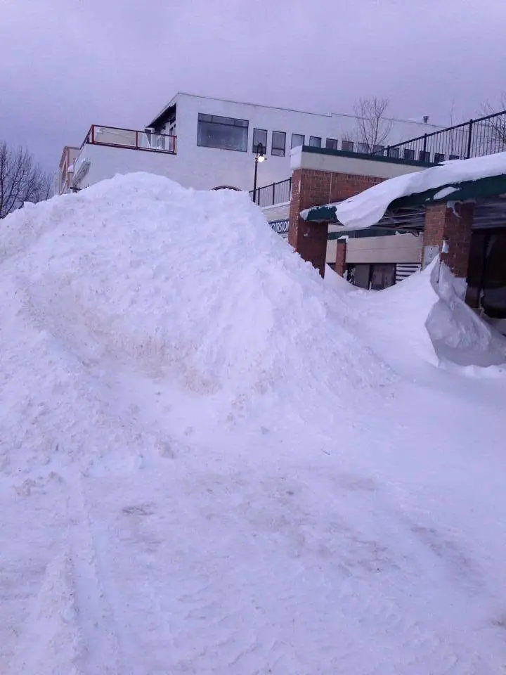 A huge snowbank rises in front of a multi-level building with balconies, brick pillars, and a green awning.