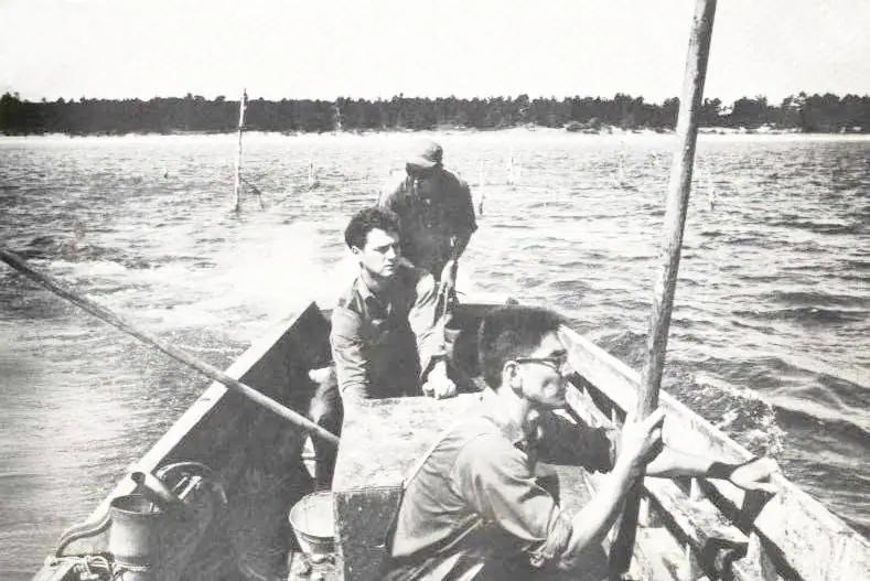 Three men sit in a small wooden boat on open water, with poles and buckets visible around them.