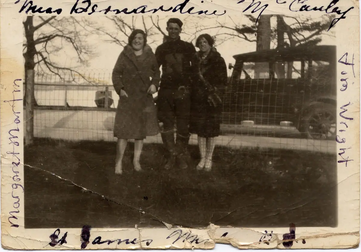 Three adults pose outdoors behind a wire fence, with a building and automobile in the background.