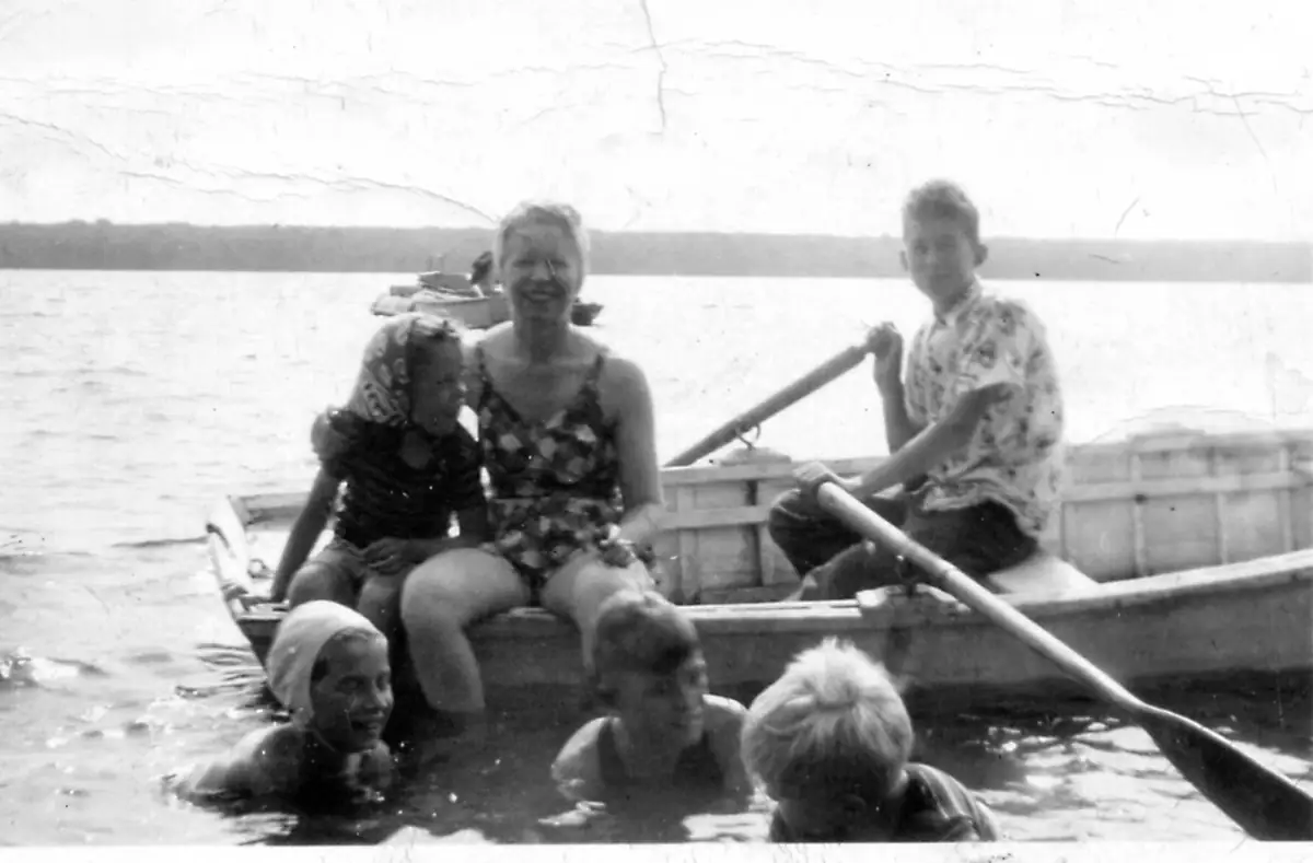 Adults and children gather in a rowboat while several children swim beside it.