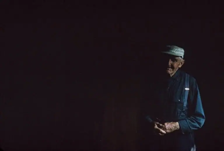 Portrait of an older man in a cap and blue shirt holding a small object against a dark background.