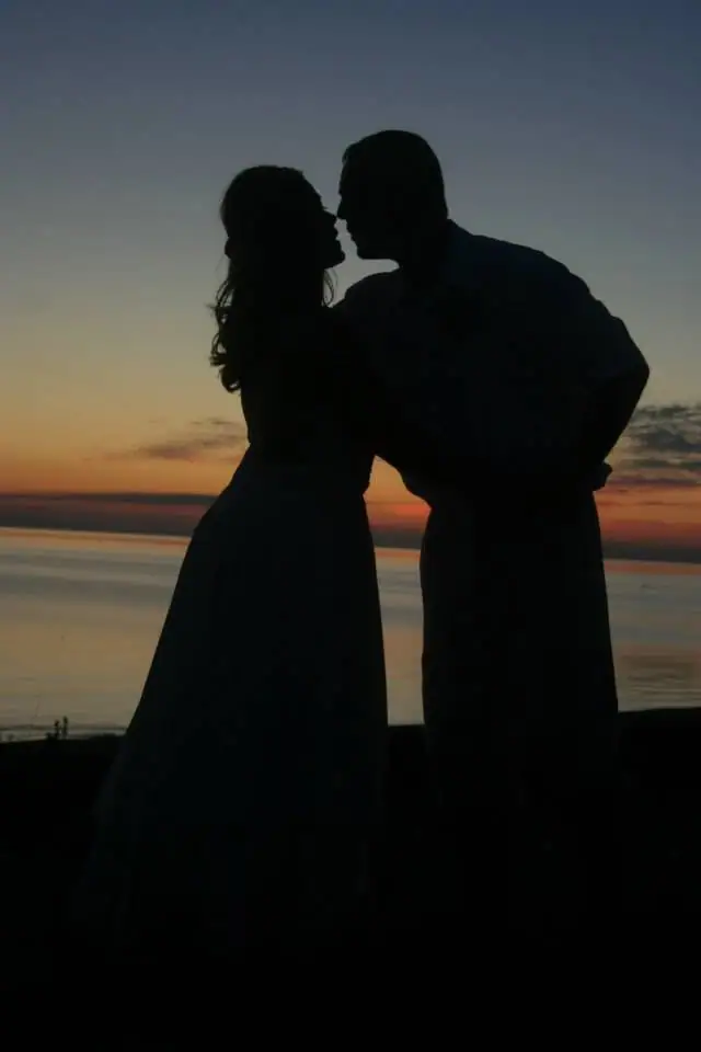 Two silhouetted people face each other against a glowing sunset over calm water.
