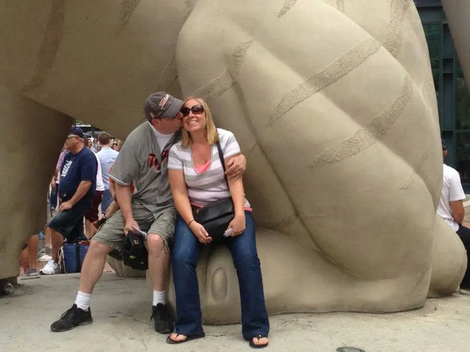 Two adults pose beside a large tan abstract sculpture while other people gather behind them.