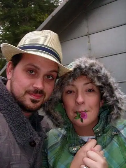 Two adults pose closely outdoors, wearing hats and plaid jackets, while one holds a pink flower in her mouth.
