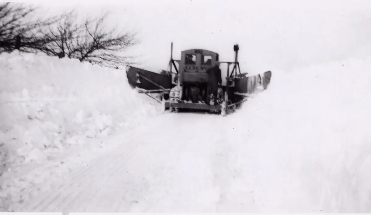 A heavy snowplow clears a snow-covered road between high snowbanks.
