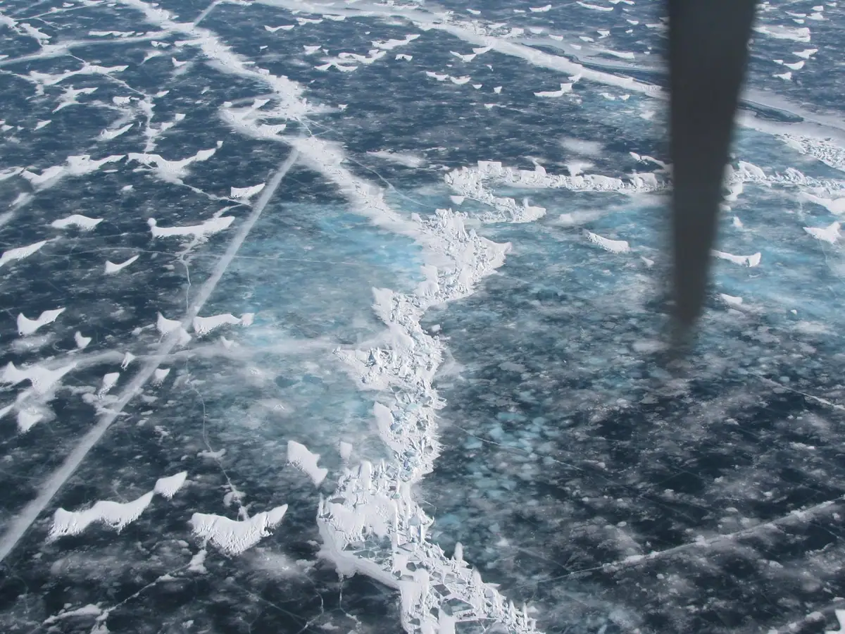 Aerial view of broken ice with a prominent winding pressure ridge and a dark vertical obstruction.