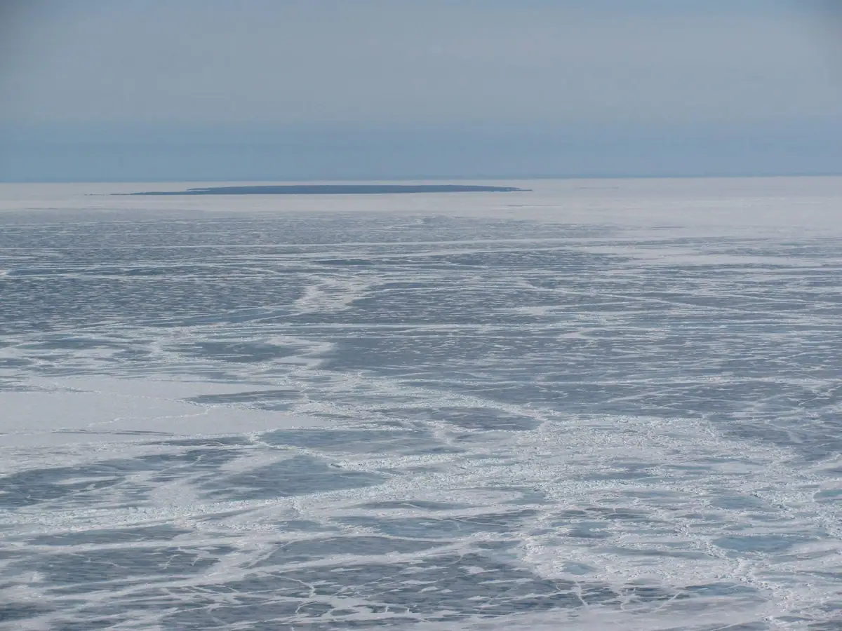 A broad frozen expanse stretches toward a low dark landform beneath a pale blue sky.