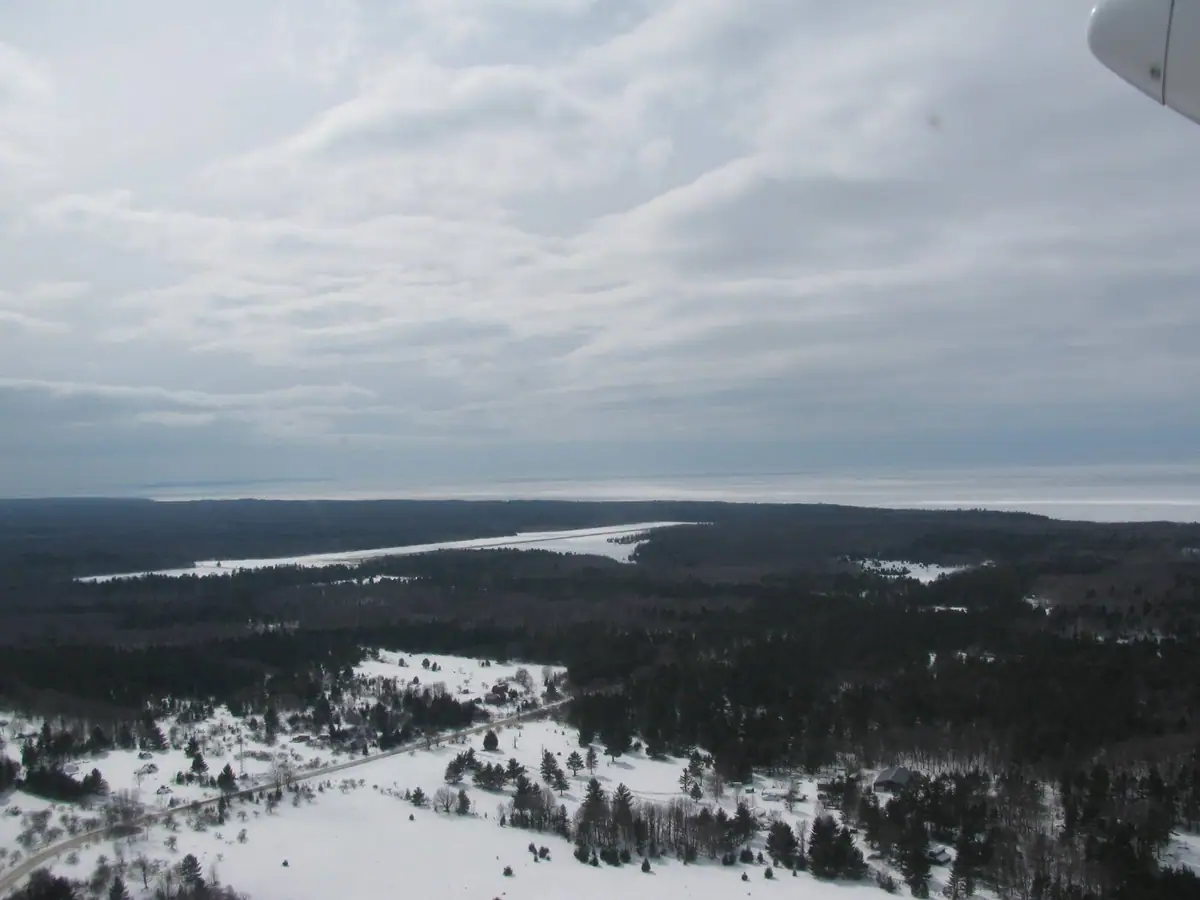 Aerial winter view of snowy fields, forest, shoreline, and water beneath an overcast sky.