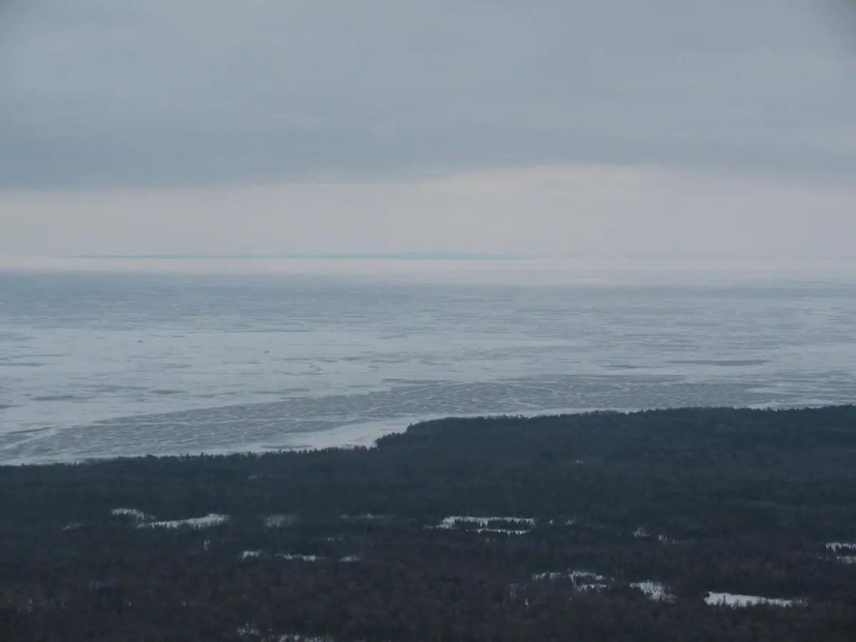 A snow-dusted forest borders broad, partly frozen water beneath a gray, overcast sky.