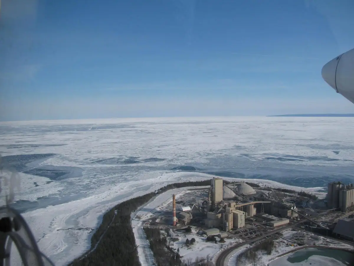 Aerial view of an industrial plant beside snow-covered ice and a forested shoreline.