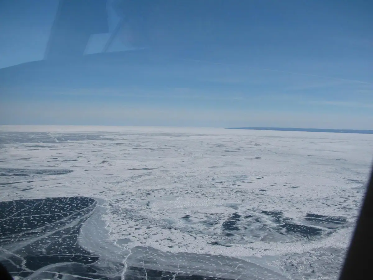 Aerial view of broad frozen water with fractured ice, dark openings, and a distant low horizon.