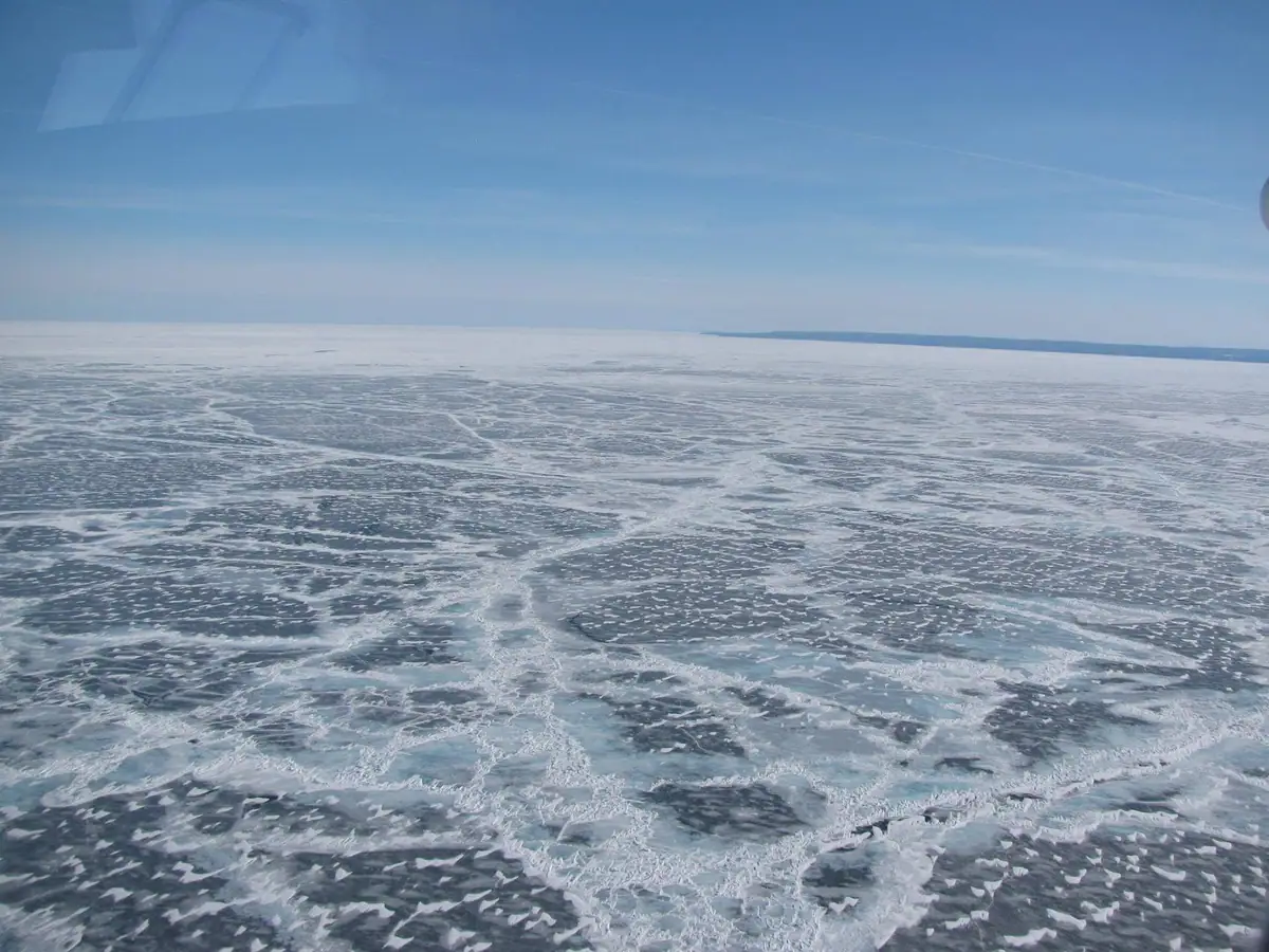 Aerial view of cracked ice stretching toward a distant shoreline beneath a clear blue sky.
