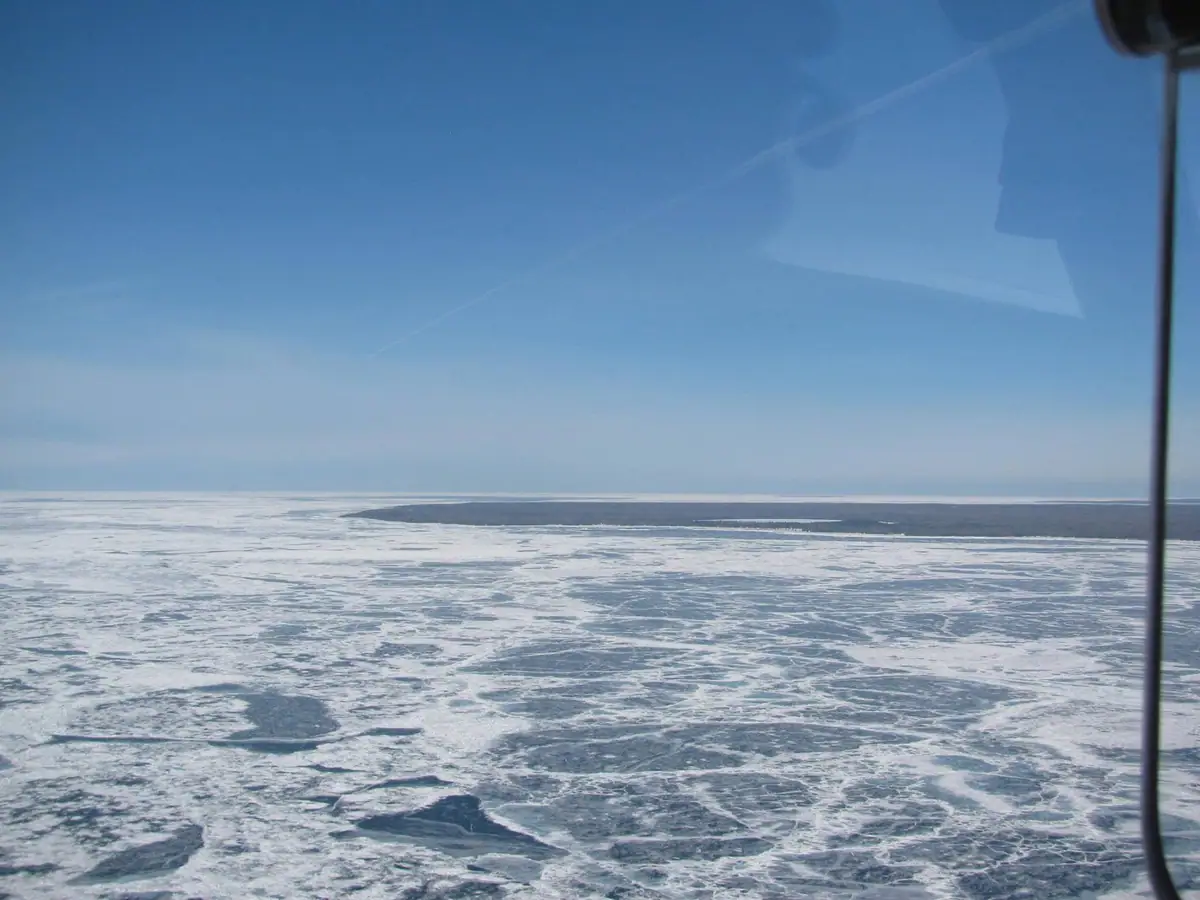 Aerial photograph of ice-covered water and a dark strip of land beneath a clear blue sky.