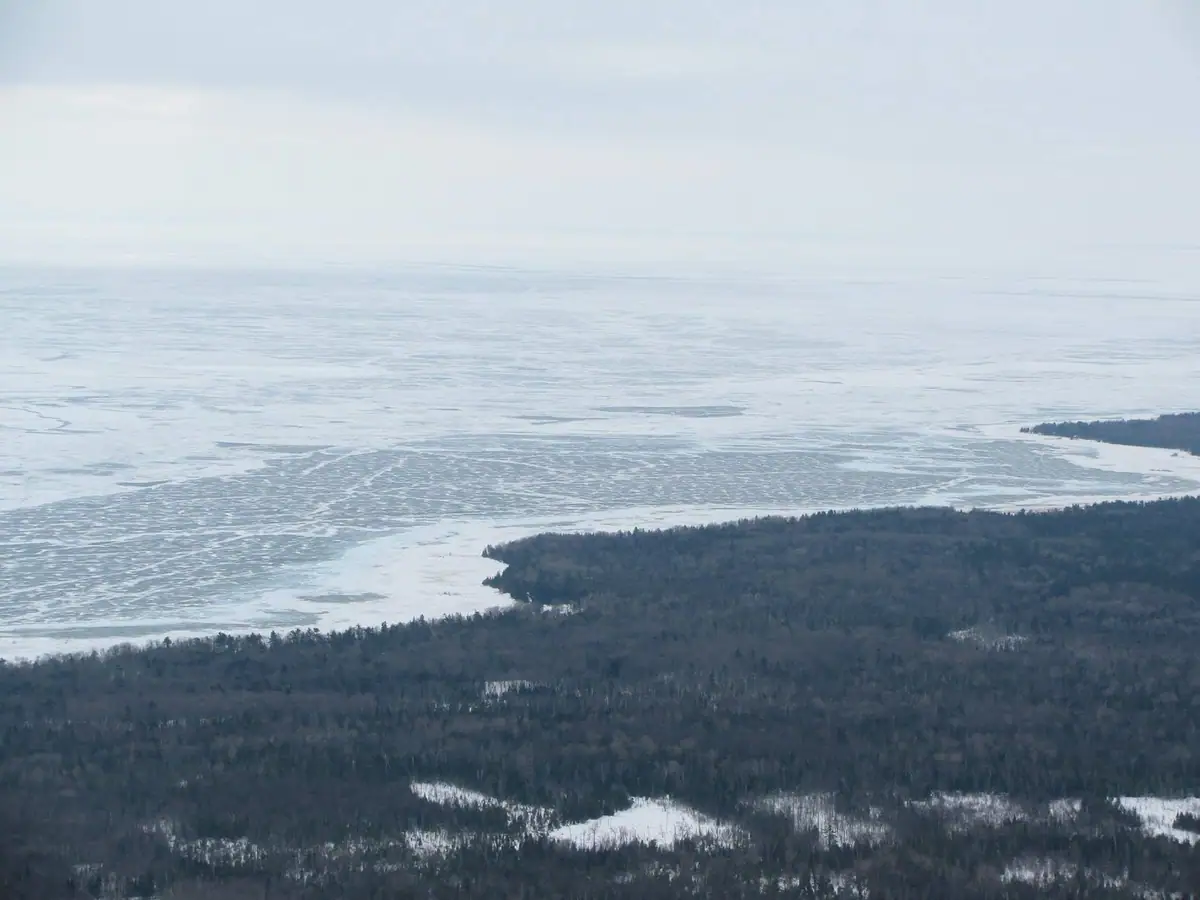 Aerial view of ice-covered water beside a wooded shoreline in winter.