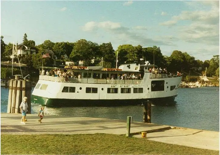 White passenger ferry carrying people beside a pier, with an American flag and wooded waterfront behind it.