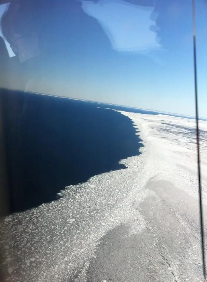 A dark expanse of open water borders bright, broken ice beneath a blue sky, seen through a window.