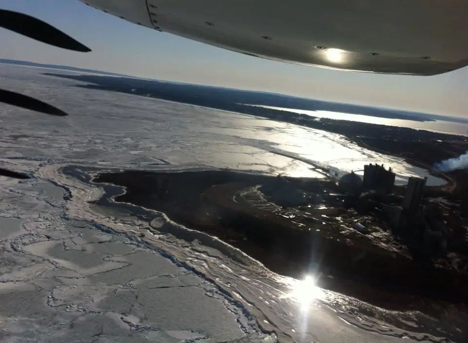Aerial view of ice-covered water and an industrial facility photographed through an aircraft window.