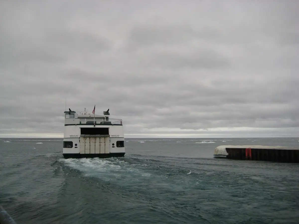 A white ferry leaves a dock, creating a wake beneath a dark, overcast sky.