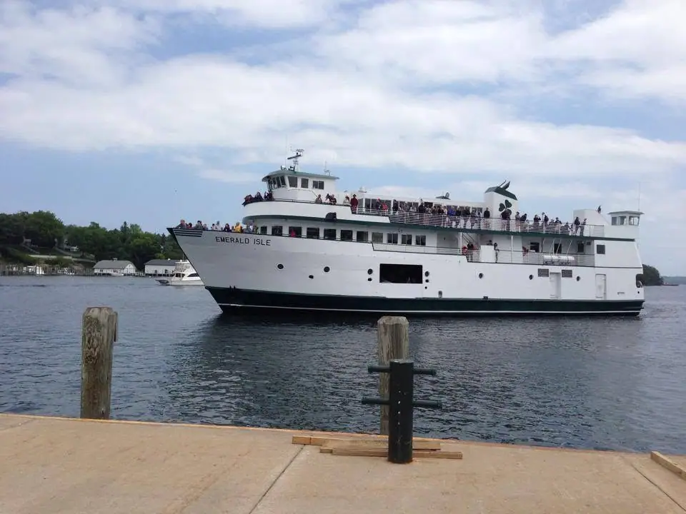 A crowded white ferry with green trim passes a concrete dock beneath a partly cloudy sky.
