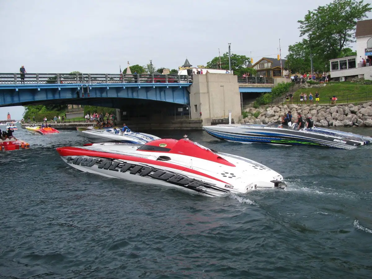 Several colorful speedboats pass beneath a blue bridge while spectators watch from the bridge and rocky shore.