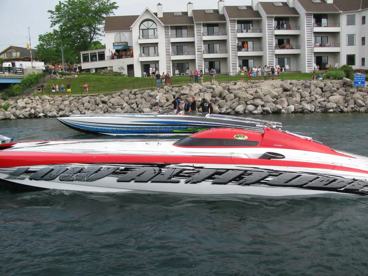 Two brightly colored speedboats pass spectators along a rocky waterfront.