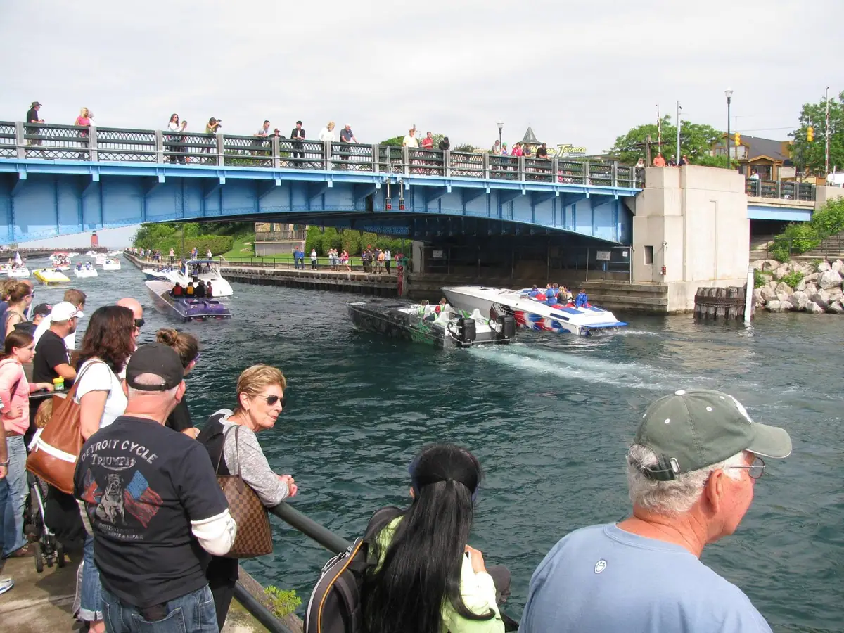 Spectators watch several motorboats pass beneath a blue steel bridge.