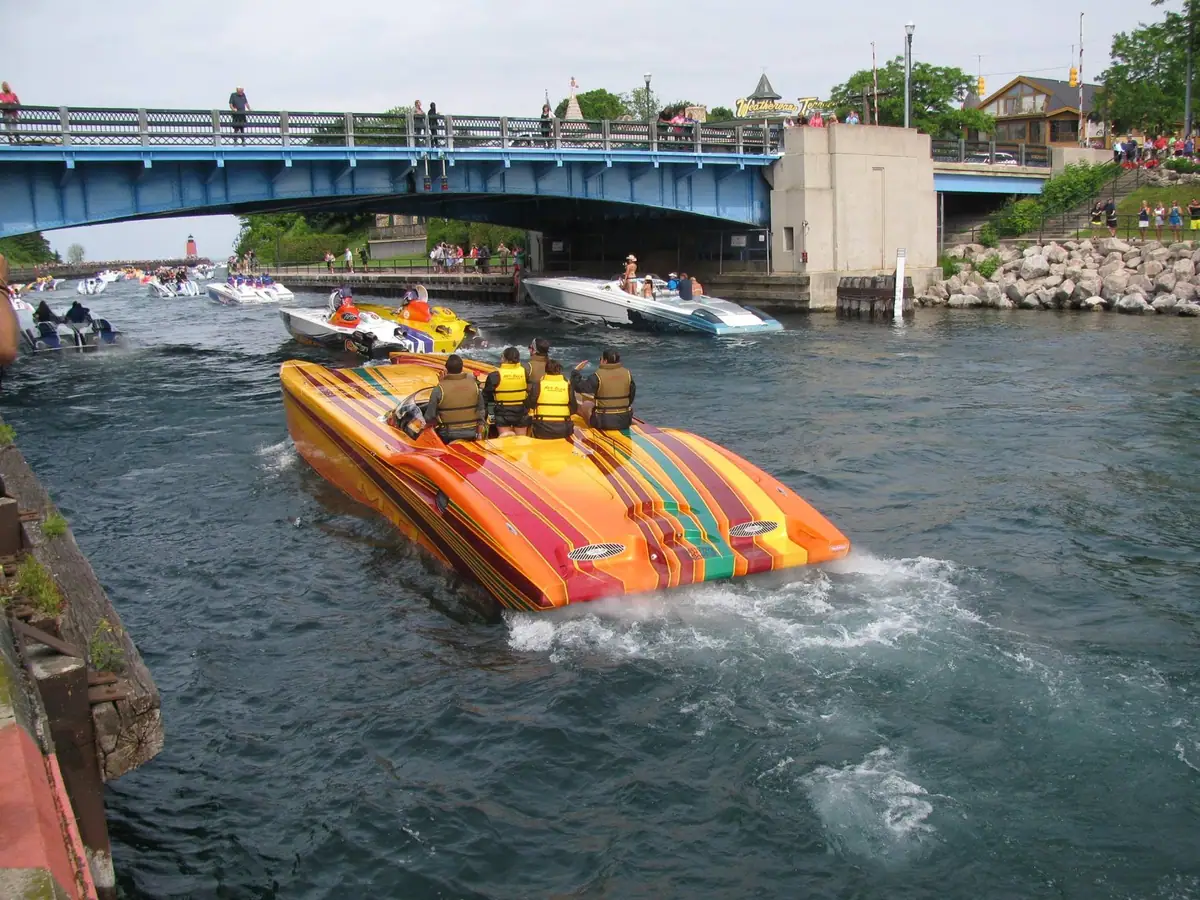 Colorful speedboats move through a channel beneath a bridge as spectators watch from the bridge and shoreline.