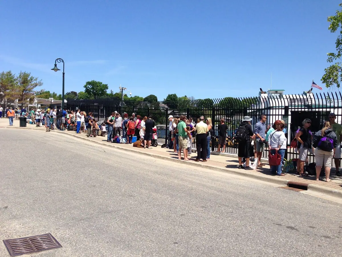 A long line of people waits beside a black metal fence along a sunny street.