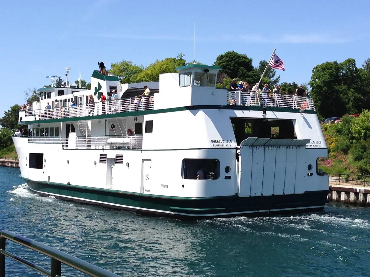 A white-and-green passenger ferry carries people past a dock, with an American flag flying above its upper deck.