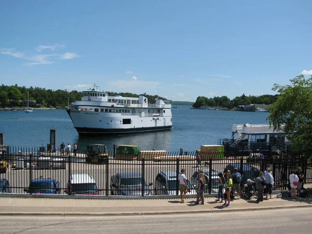 A white passenger ferry approaches a waterfront pier as people gather beside a fenced sidewalk.