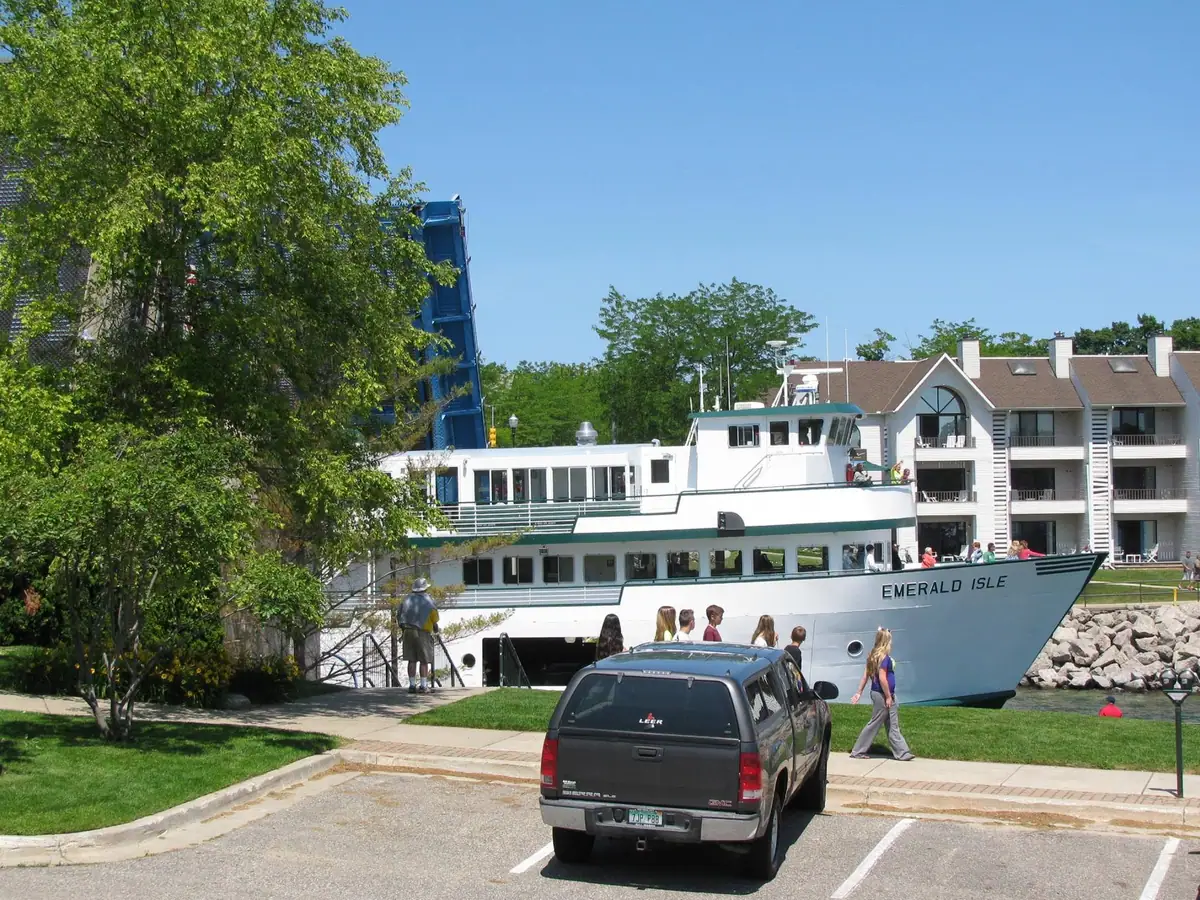 White passenger ferry beside a pier, with people, a parked SUV, trees, and a blue lift structure.