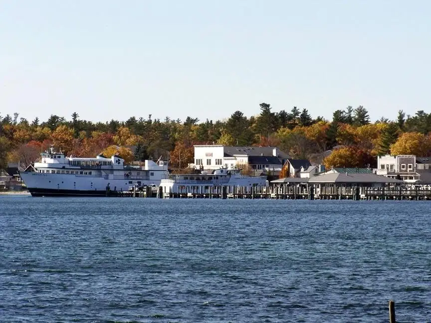 White ferry moored beside a long pier with autumn-colored trees and waterfront buildings behind it.