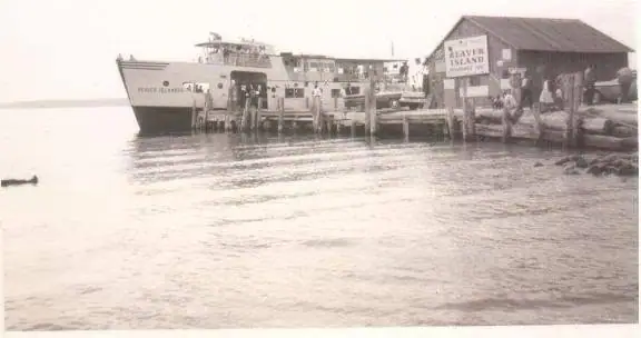A ferry is moored beside a wooden pier and shed, with people gathered along the waterfront.