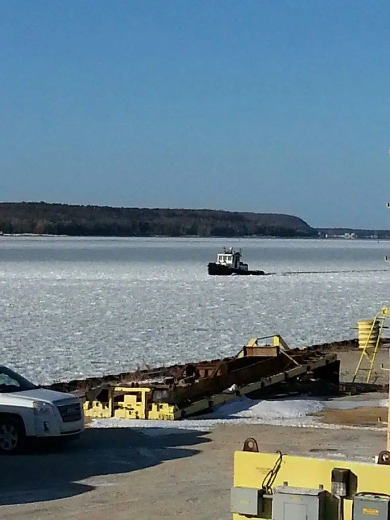 A small tugboat breaks through ice near shore while yellow heavy equipment and a car sit in the foreground.