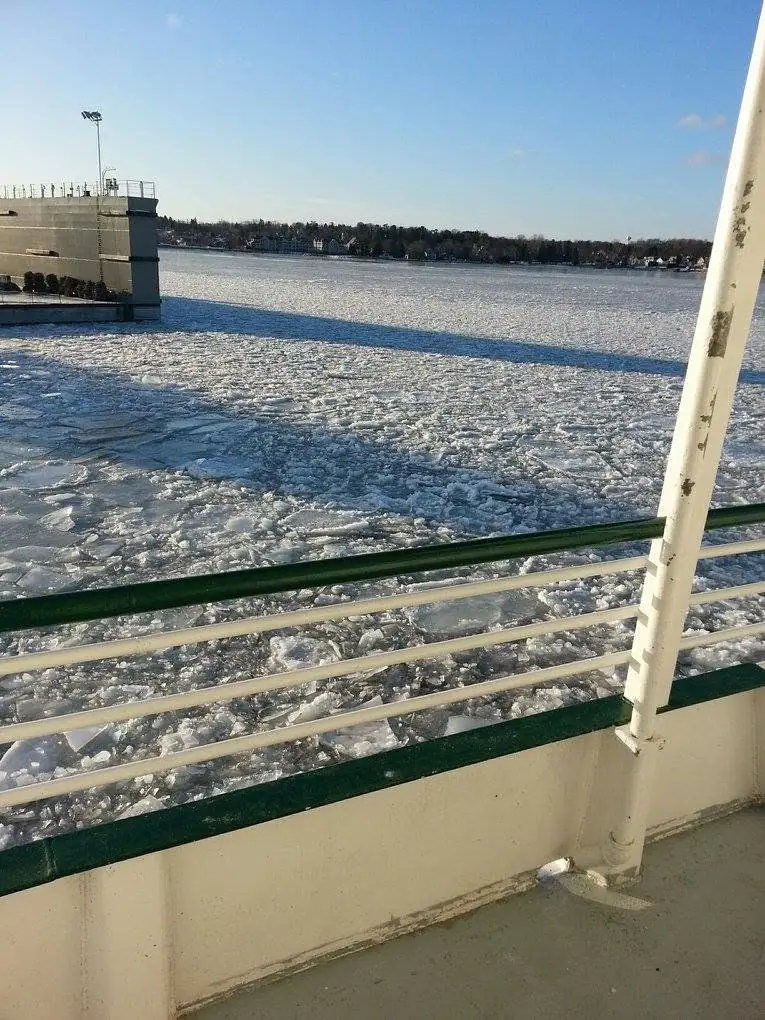 View from a boat deck across ice-covered water toward a concrete waterfront structure and wooded shoreline.