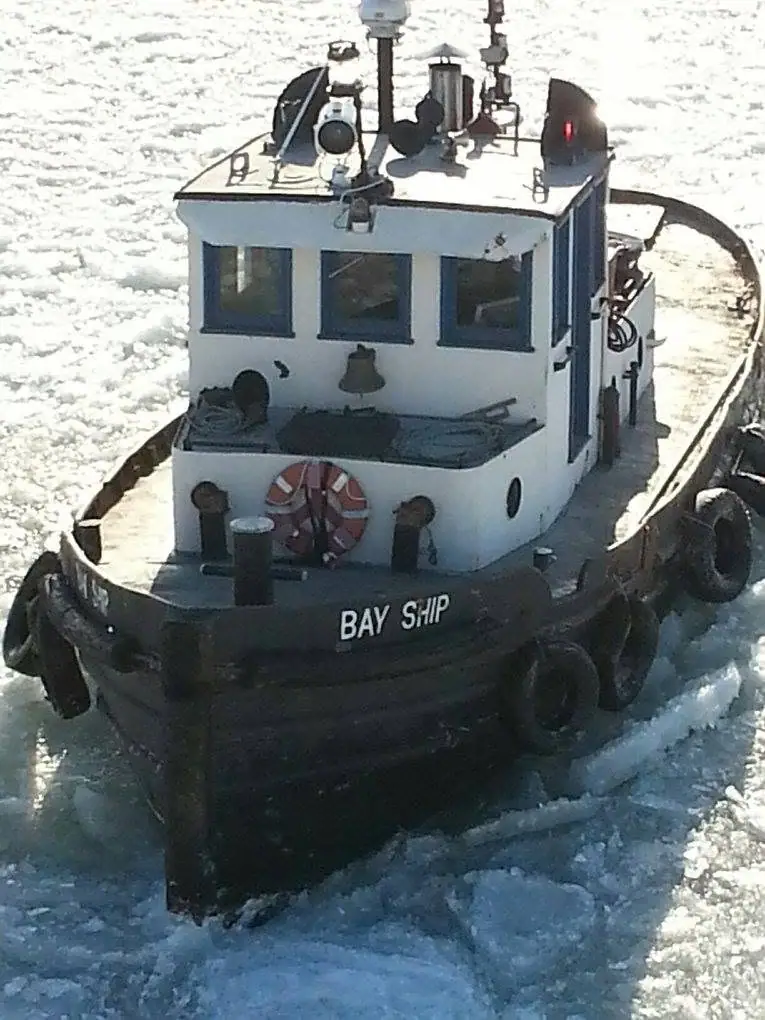 A small tugboat pushes through ice, with “BAY SHIP” painted on its dark hull.