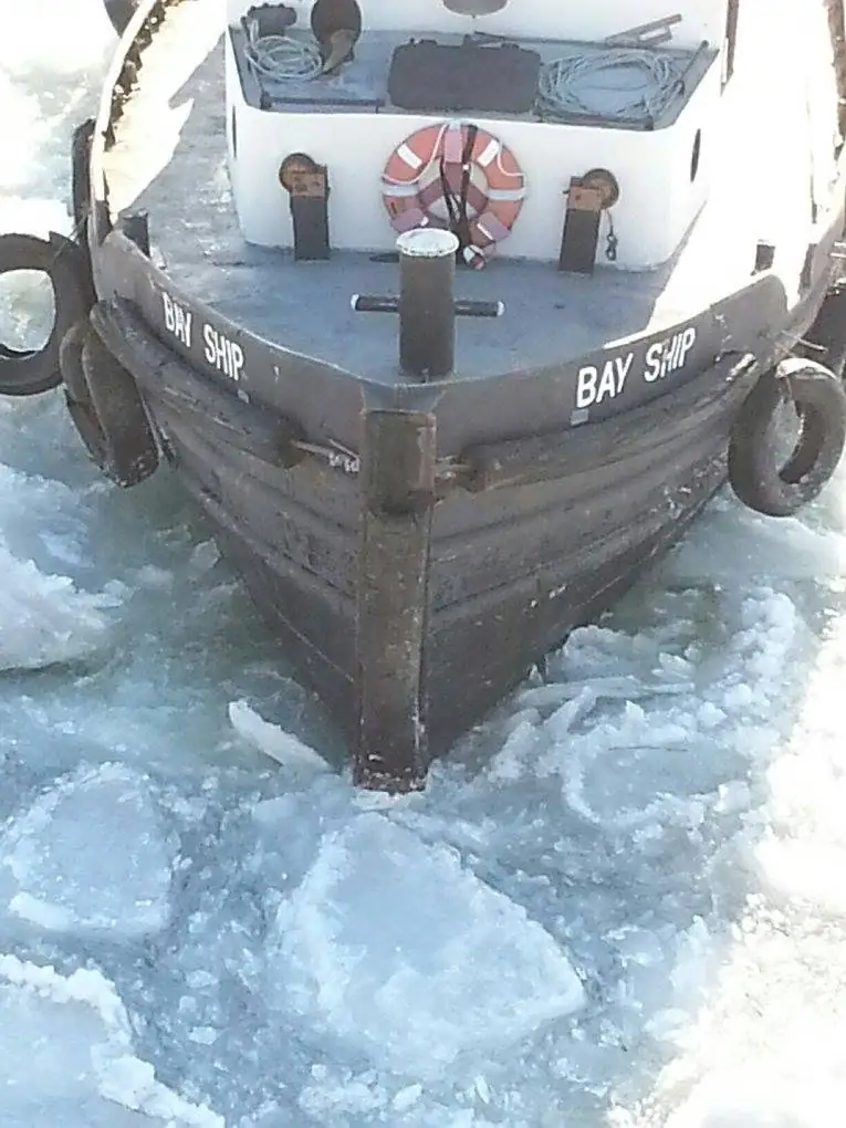 A dark-hulled boat marked BAY SHIP pushes through broken ice, with a life ring and ropes on its deck.