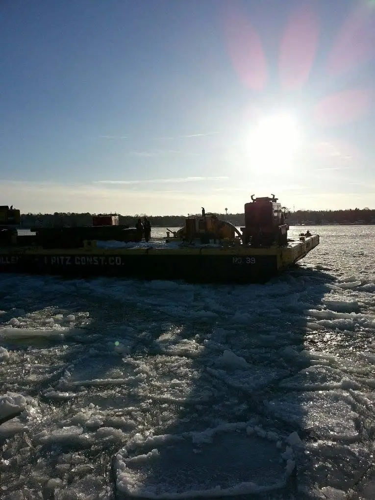 A flat-deck barge marked “PITZ CONST. CO.” and “NO. 39” pushes through broken ice in bright sunlight.