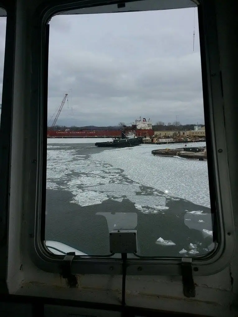 A view through a vessel window toward a freighter, tugboat, dock, and floating ice.