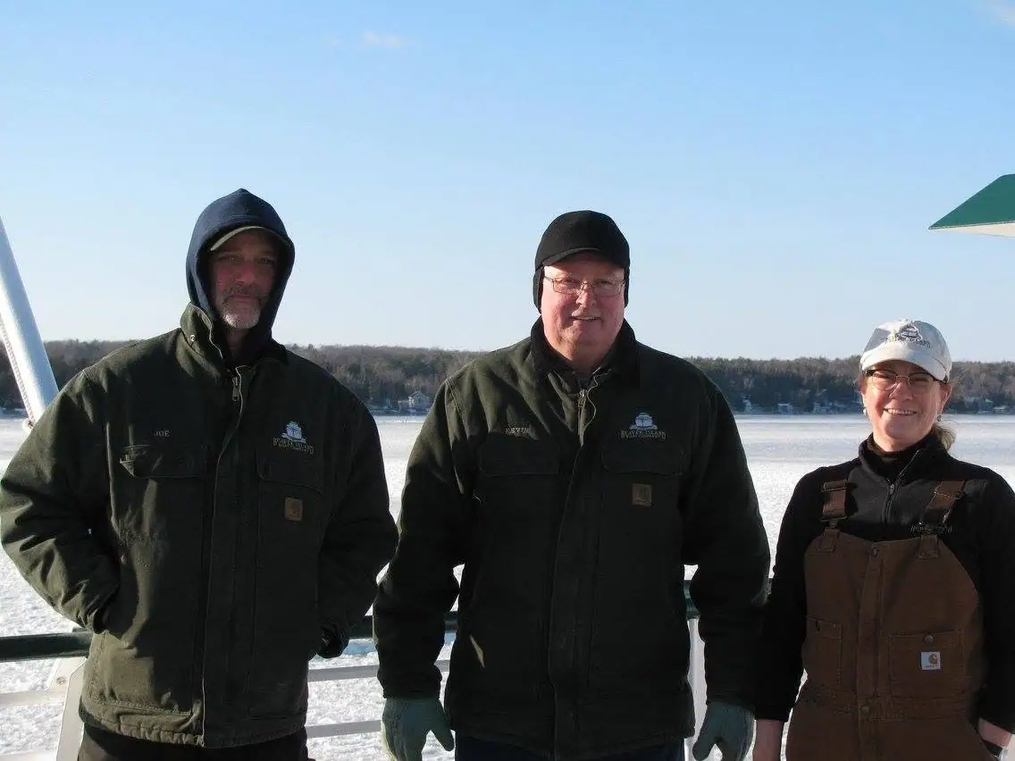 Three adults in winter work clothing stand on a boat deck beside an ice-covered expanse.
