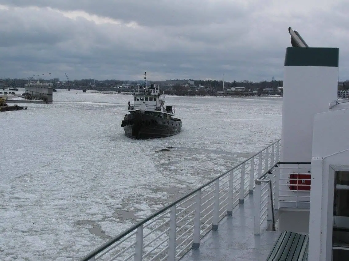 A tugboat moves through broken ice beside the deck of a larger vessel.