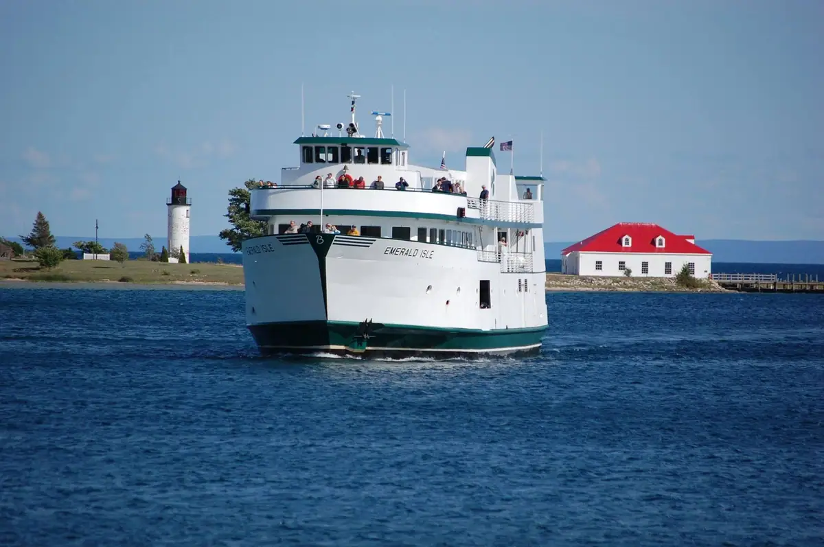 A white-and-green passenger ferry approaches a shoreline with a lighthouse and red-roofed building.