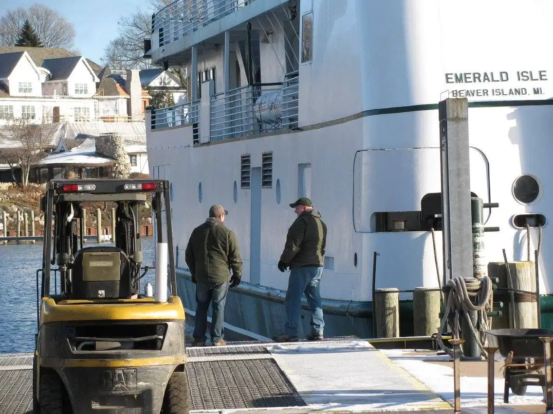 Two dock workers stand beside a white ferry and yellow forklift at a waterfront loading ramp.
