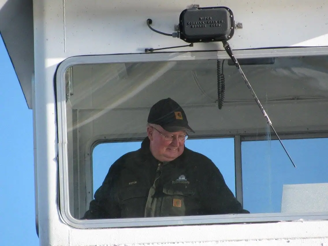 A man in a cap and glasses looks down from inside a boat wheelhouse beneath a windshield-wiper motor.