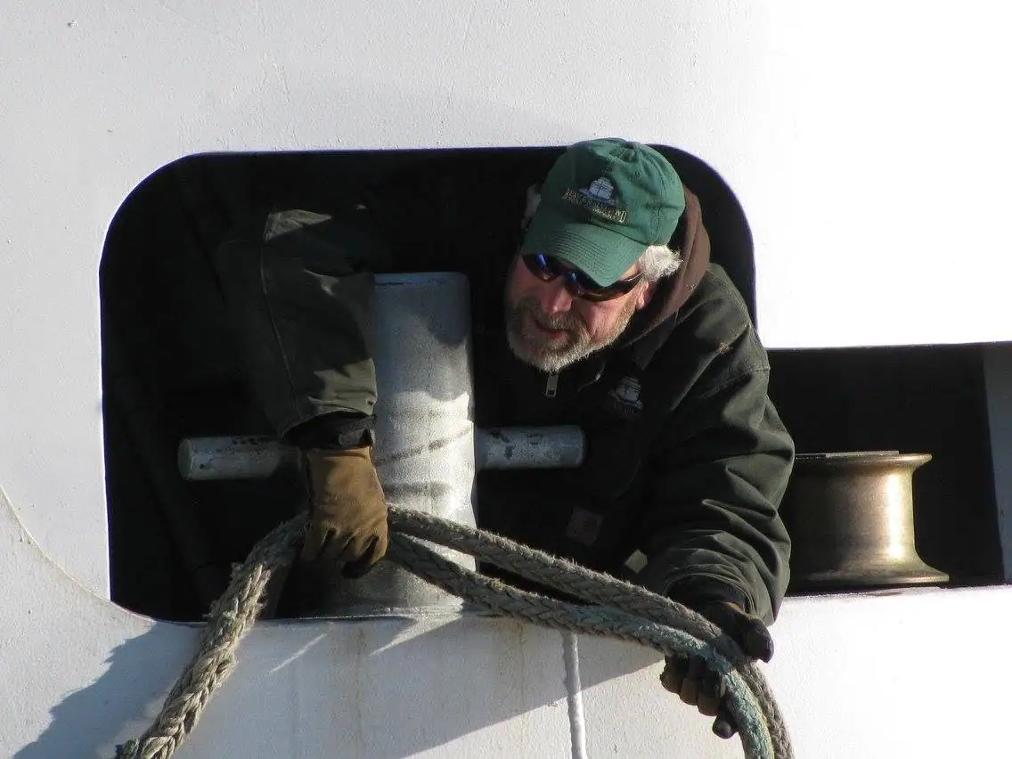 A bearded man in sunglasses handles a thick rope beside a metal winch on a vessel.