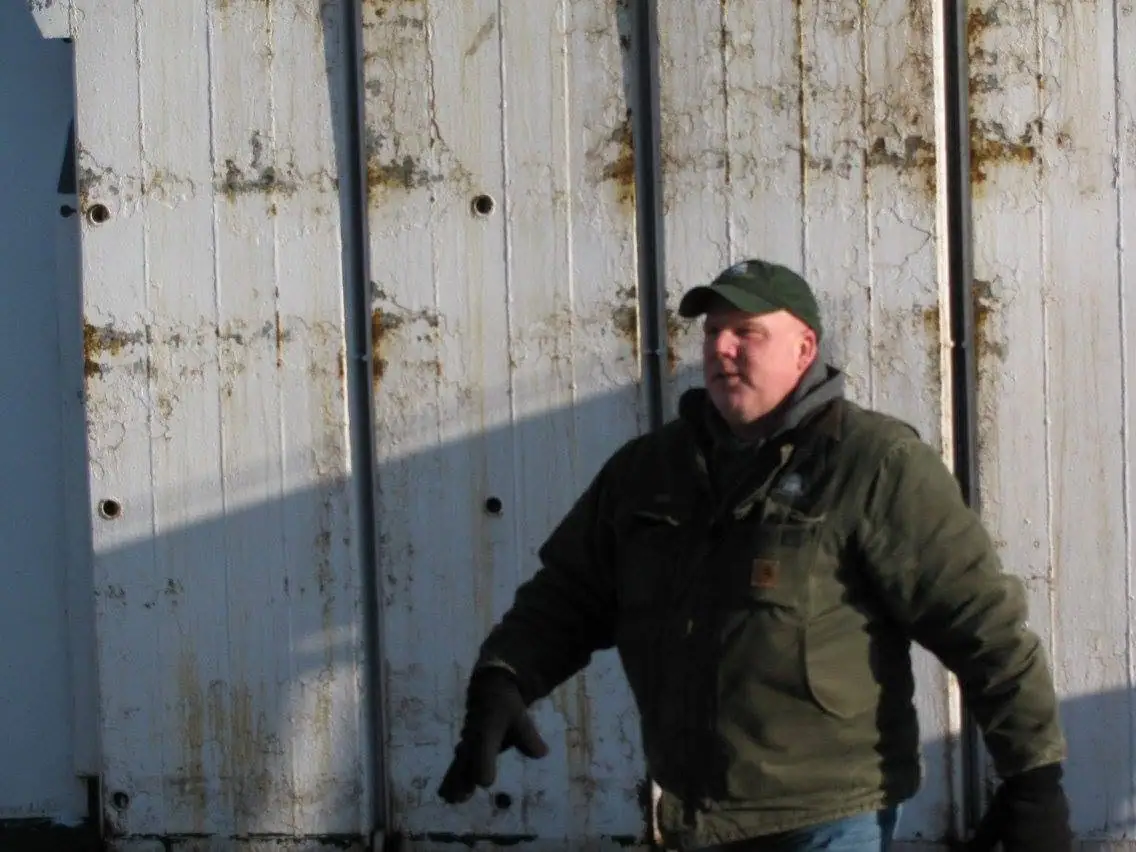 A man in a green cap and winter jacket stands before a weathered, rust-streaked metal wall.