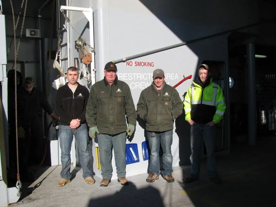 Five men in work clothing pose on a vessel deck beside machinery and crew-only warning signs.