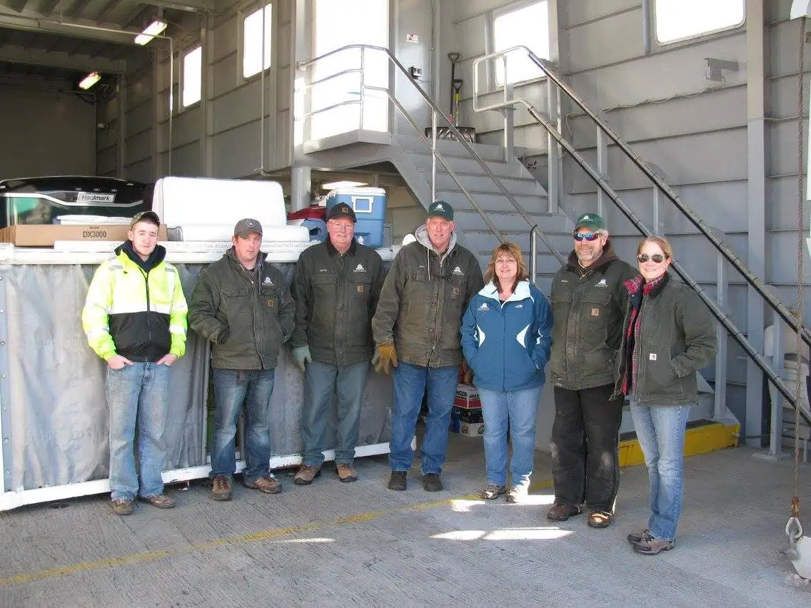 Seven people pose beside a white Haulmark trailer inside a vessel with metal stairs and railings.